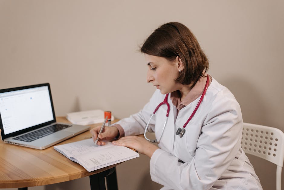 A female doctor in a lab coat writing a prescription in a clinic office setting with a laptop on the table.