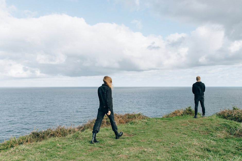 Two people in dark clothing stand on a grassy cliff edge overlooking the ocean under a cloudy sky.