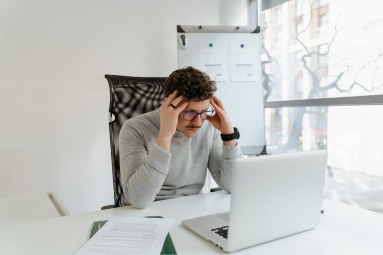 Young man concentrating on work at a desk with laptop in modern office setting.