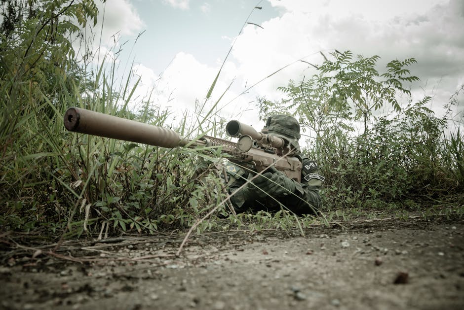 A camouflaged sniper with rifle hidden in grasslands during daytime surveillance.