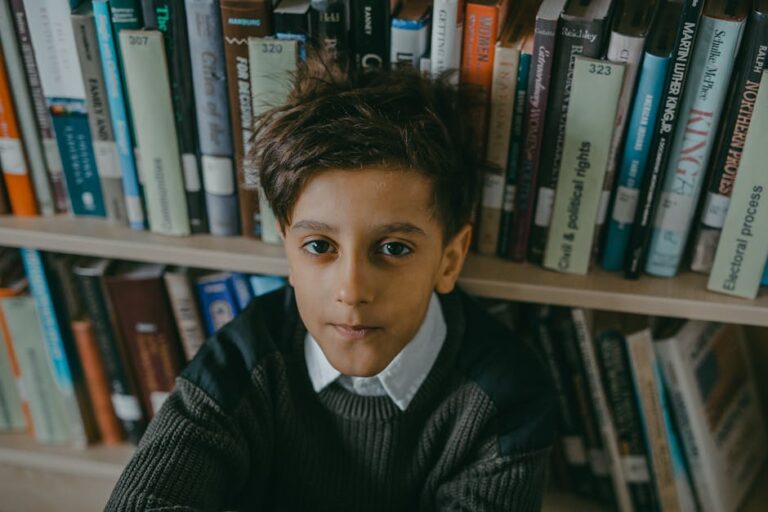 A young boy sits pensively against a bookshelf lined with various books.