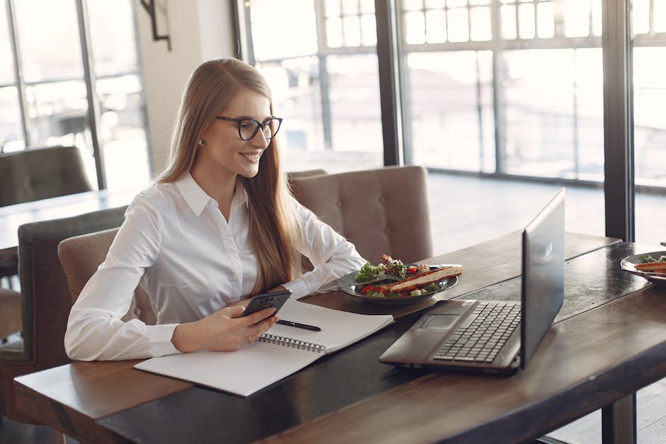 Smiling businesswoman in white shirt using smartphone and laptop at a café while enjoying lunch.