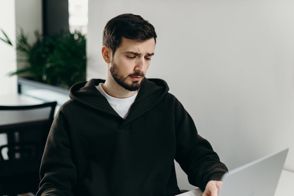 A young man in a hoodie working on a laptop at a home office desk, focused and thoughtful.