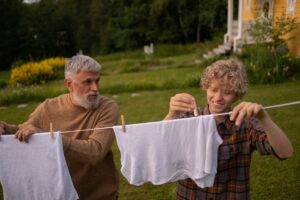 Elderly man and teenage boy joyfully hanging laundry in a sunny garden setting.