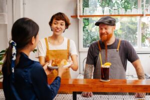 Cheerful female worker passing delicious puff to unrecognizable partner above counter near smiling tattooed man