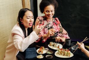 Two women smiling and taking photos while enjoying sushi at a Japanese restaurant.