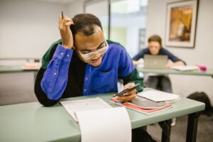 A college student studying in class using a smartphone, with papers and books on the desk.